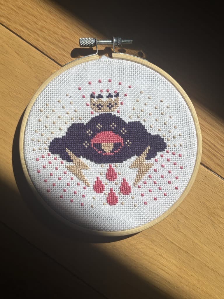 A finished cross-stitch of a crowned rain cloud in a wooden hoop