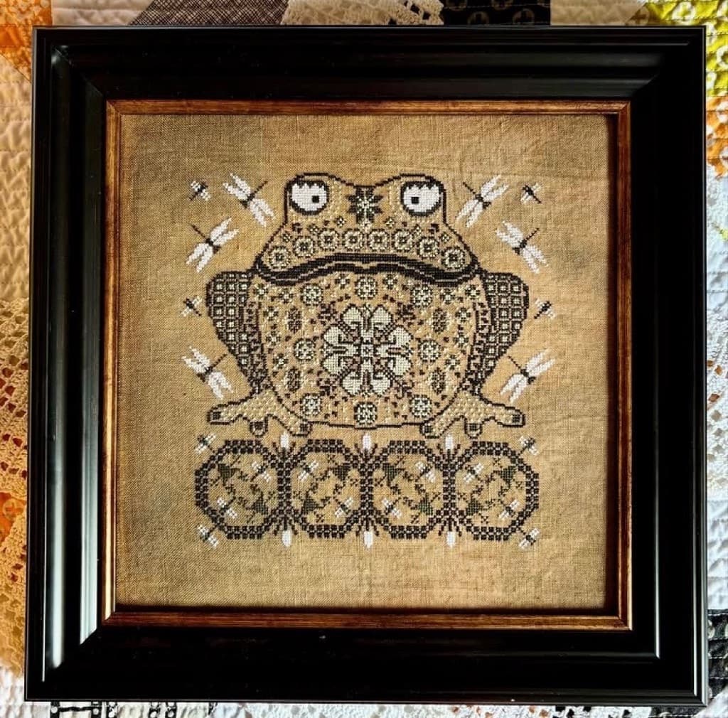 A framed cross-stitch of an ornate toad, ringed by dragonflies
